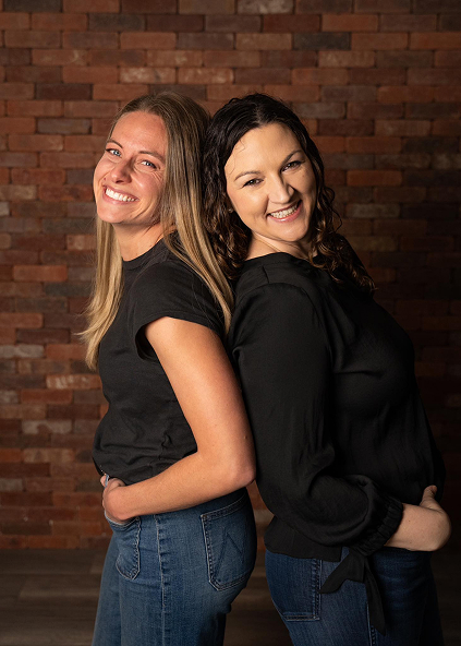 Golden Hill Family Dentistry | Two women standing back-to-back smiling in front of a brick wall.